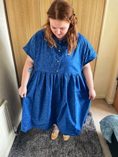 A woman with loose wavy auburn hair is looking towards the floor, wearing a blue shirt dress with gathered skirt.